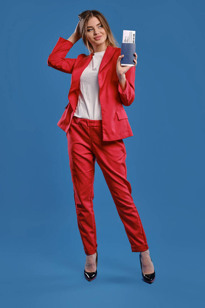 Blonde girl in white blouse, red pantsuit, high black heels. She touching hair, holding passport and ticket, posing on blue background. Full length - Photo, Image