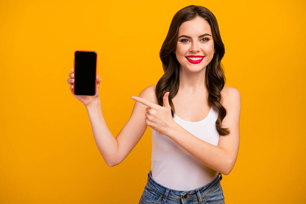 Photo of pretty amazing lady hold telephone hand indicating finger touch screen advertising new gadget model wear white tank-top jeans isolated bright yellow color background - Foto, Imagem