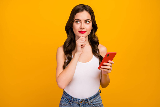 Photo of pretty lady hold telephone hands look dreamy up empty space think over new post idea arm on chin wear white tank-top jeans isolated bright yellow color background - Photo, Image
