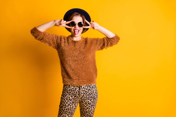 Foto von schönen lustigen Dame halten Finger V-Zeichen Symbol in der Nähe der Augen gute Laune Stick Zunge aus Mund tragen Pullover Leopardenhosen Sonnen Spezifikationen Retro-Kappe isoliert gelbe Farbe Hintergrund - Foto, Bild