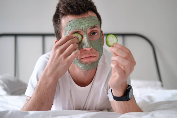 Młody mężczyzna w białej koszuli z zieloną maską kosmetyczną trzymający kawałki ogórka - Zdjęcie, obraz