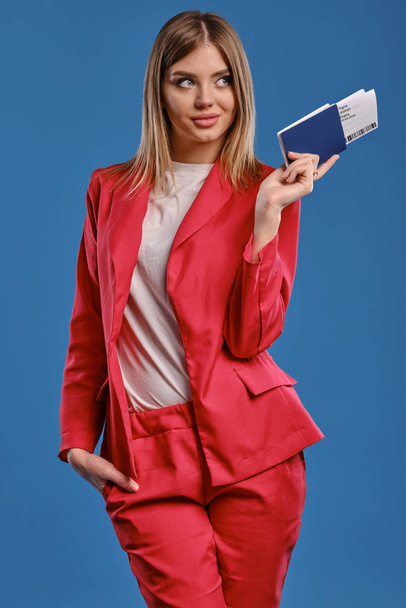 Blonde girl in white blouse and red pantsuit. She smiling, holding passport and ticket while posing on blue studio background. Close-up - Φωτογραφία, εικόνα
