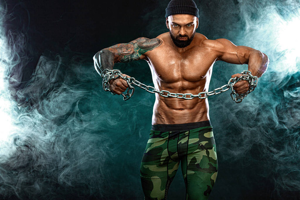 Athlete bodybuilder on black background with lights and smoke. Men fashion. Portrait of a brutal bearded man topless with chains. - Foto, imagen
