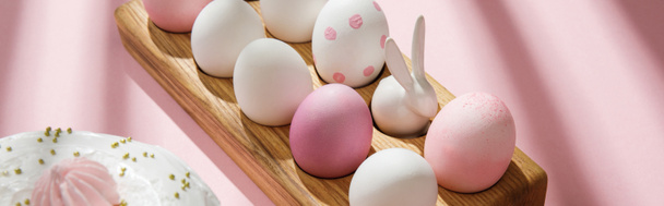Huevos de Pascua y conejito decorativo en tablero de madera cerca de la torta sobre fondo rosa, tiro panorámico
 - Foto, Imagen