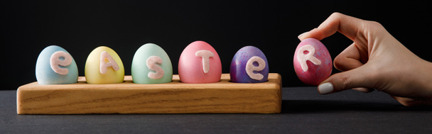 Cropped view of woman holding painted chicken egg near board with colorful eggs with easter lettering on grey and black, panoramic shot - Photo, Image