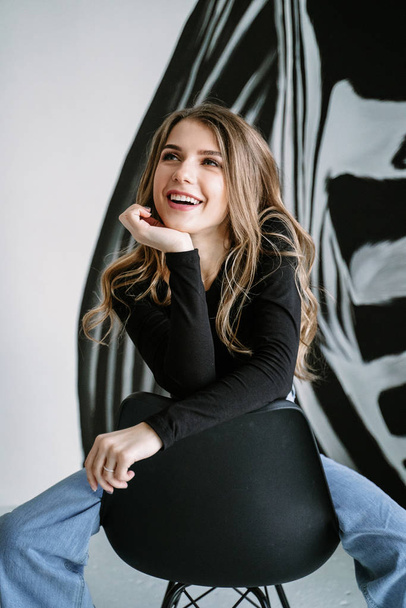 Portrait of a beautiful girl with blond long hair in a black T-shirt and jeans sitting on a black chair against the background of a wall with a zebra pattern - Фото, изображение
