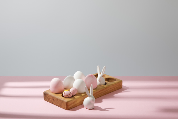 Huevos de Pascua sobre tabla de madera con conejos decorativos sobre fondo rosa y gris
 - Foto, Imagen