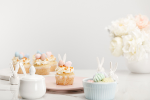 Selective focus of cupcakes, decorative bunnies, sugar bowl, meringues and vase with flowers on grey background - Photo, Image