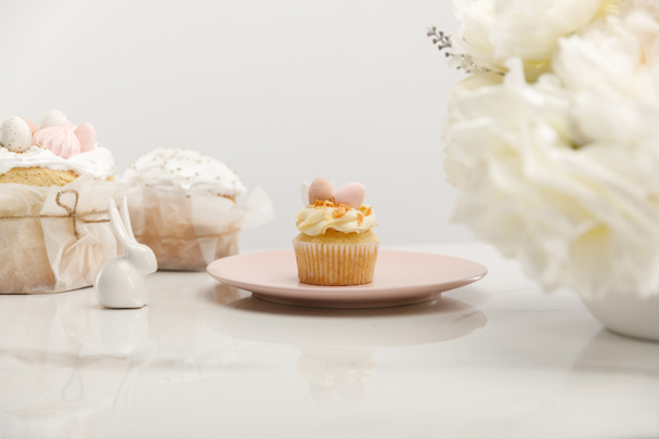 Selective focus of cupcake on plate, decorative bunnies near vase with bouquet and easter cakes isolated on grey - 写真・画像