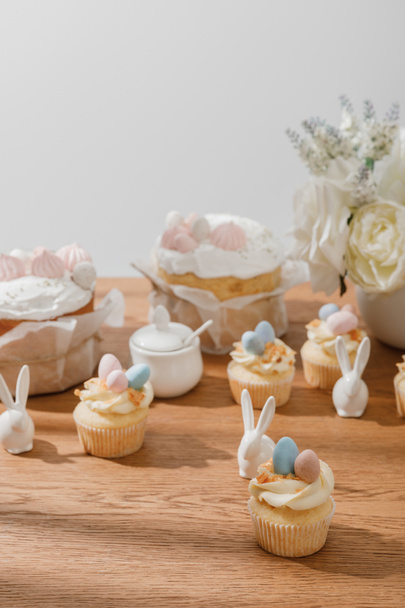 Selective focus of cupcakes with decorative rabbits, sugar bowl, easter cakes and vase with bouquet isolated on grey - Photo, Image