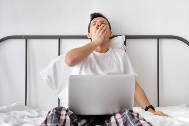 Sonriente joven guapo en una camisa blanca y pijama sentado en una cama con ordenador portátil y estiramiento
 - Foto, imagen