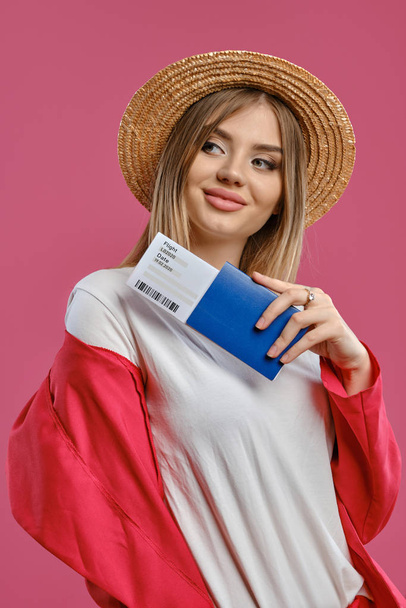 Mujer rubia con sombrero de paja, blusa blanca y pantalón rojo. Ella sonrió, sosteniendo pasaporte y boleto mientras posaba sobre fondo rosa del estudio. Primer plano
 - Foto, Imagen