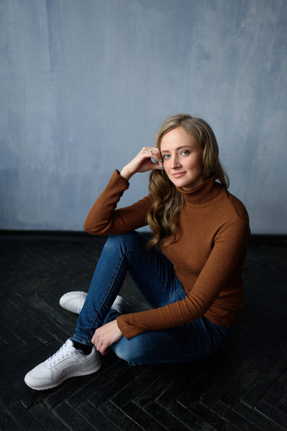 Young beautiful woman with long straight blonde hair in blue jeans and bright blouse sitting on black wooden table with crossed legs and looking in camera. - Фото, изображение