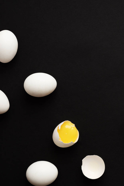 White eggs lie scattered on a black background. One egg is broken and the yolk is visible. Minimalistic design and color trend 2020. Top view. - Fotografie, Obrázek