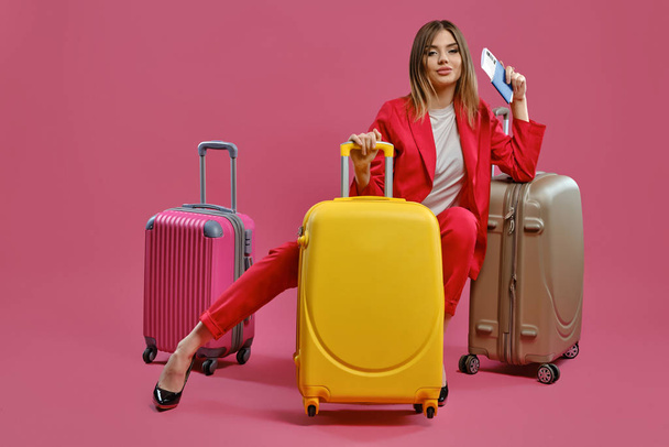 Blonde lady in red pantsuit, white blouse, black heels. She smiling, sitting among colorful suitcases, holding passport and ticket, pink background - Photo, Image