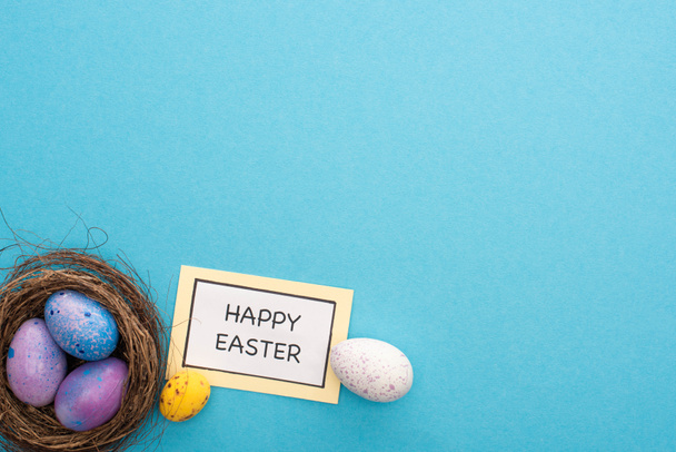 Πάνω άποψη της φωλιάς με πασχαλινά αυγά κοντά σε κάρτα με γράμματα Καλό Πάσχα σε μπλε φόντο - Φωτογραφία, εικόνα