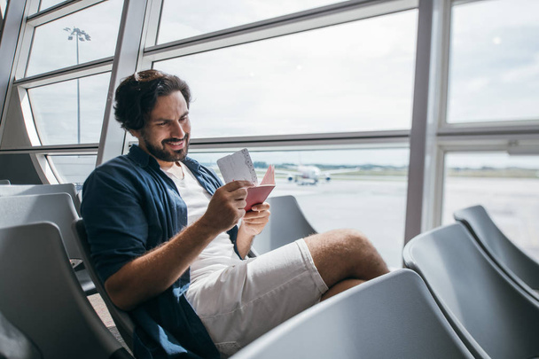 A man sits in a waiting room at the gate at the airport. Young guy, a traveler with a backpack waiting for the departure of the plane on the background of a large window and the runway - Foto, Imagen