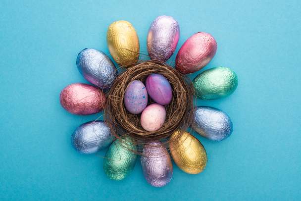 Top view of chocolate Easter eggs in colorful foil around nest with quail eggs on blue background - Zdjęcie, obraz