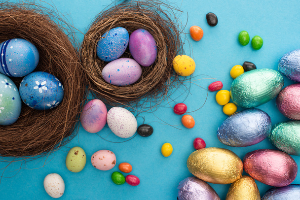 Top view of candies, chocolate Easter eggs in colorful foil near nests with painted chicken and quail eggs on blue background - Photo, Image