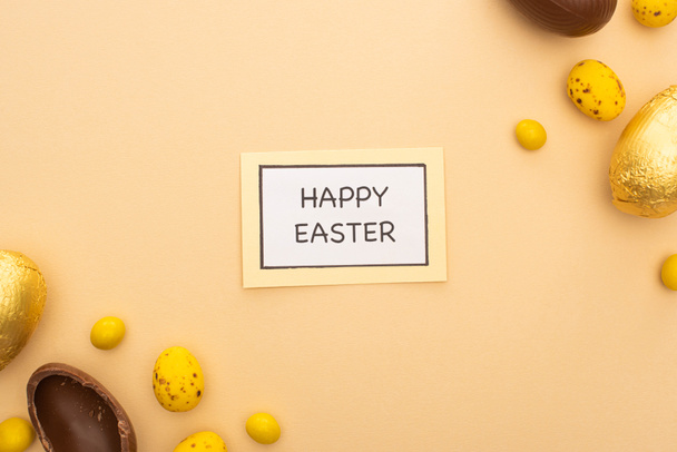 Верхний вид открытки с радостными пасхальными надписями, шоколадные яйца, конфеты и окрашенные перепелиные яйца на бежевом фоне
 - Фото, изображение