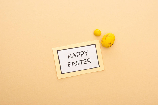 Top view of card with happy easter lettering, candy and yellow quail egg on beige background - Photo, Image
