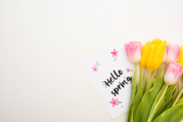 Bovenaanzicht van kaart met hallo lente belettering bij gele en roze tulpen op witte achtergrond - Foto, afbeelding