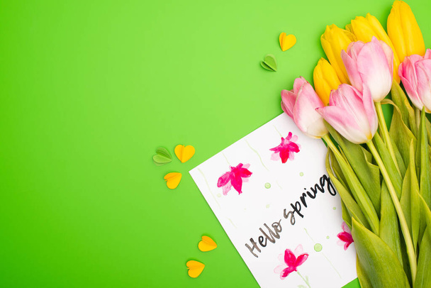 Bovenaanzicht van gele en roze tulpen, kaart met hallo lentebelettering en decoratieve hartjes op groene achtergrond - Foto, afbeelding