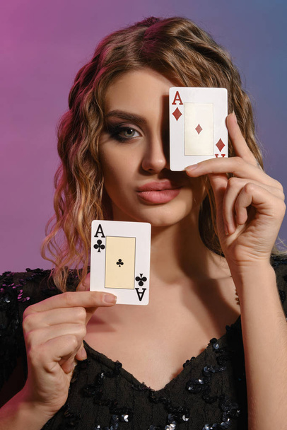 Brown-haired woman in black shiny dress showing two playing cards, posing on colorful background. Gambling entertainment, poker, casino. Close-up. - Fotoğraf, Görsel