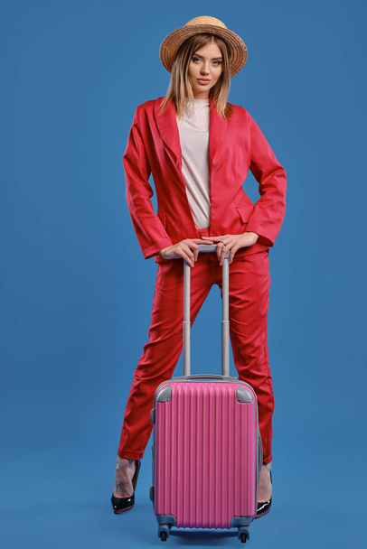 Blonde girl in straw hat, white blouse, red pantsuit, high black heels. She is leaning on a handle of pink suitcase while posing on blue background - Photo, Image