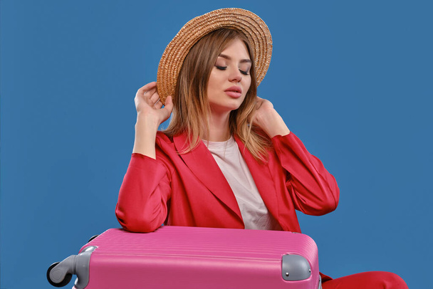 Blonde model in straw hat, white blouse, red pantsuit. Sitting leaning on pink suitcase, posing on blue background. Travelling concept. Close-up - Fotografie, Obrázek