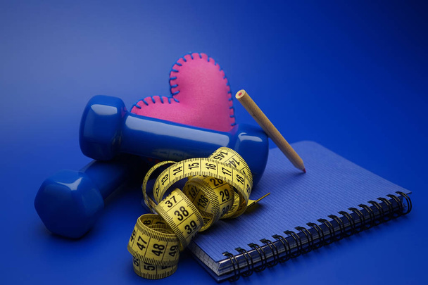 Fitness and weight loss concept with dumbbells, measuring tape, heart and new notebook on blue background - Photo, Image