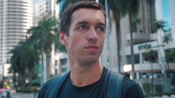 Portrait of young handsome tourist man with backpack standing outdoors alone smiling looking at camera at urban ciry background - Materiał filmowy, wideo