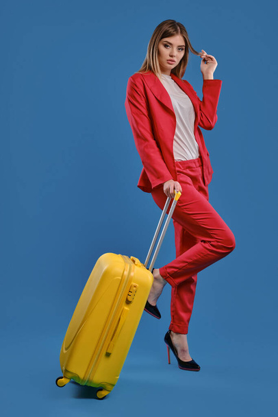 Blonde woman in red pantsuit, white blouse and high black heels. She is holding yellow suitcase, posing against blue studio background. Full length - Foto, afbeelding