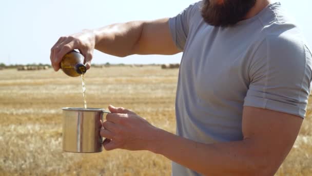 A man pours beer into a metal glass on a wheat field with bales. Fresh beer, bearded man, solar field - 映像、動画