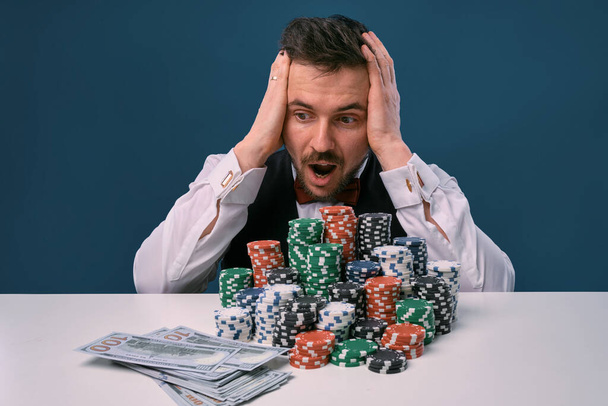 Man in black vest and shirt sitting at white table with stacks of chips and cash on it, posing on blue background. Gambling, poker, casino. Close-up. - Foto, afbeelding