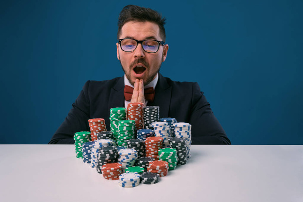 Male in glasses, black suit looking excited, sitting at white table with colored stacks of chips on it, posing on blue background. Poker, casino - Fotoğraf, Görsel