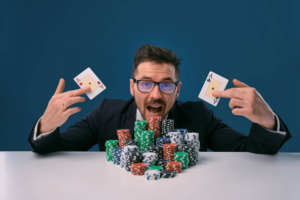 Guy in glasses, black suit sitting at white table with stacks of chips, holding two playing cards, posing on blue background. Poker, casino. Close-up. - Fotoğraf, Görsel