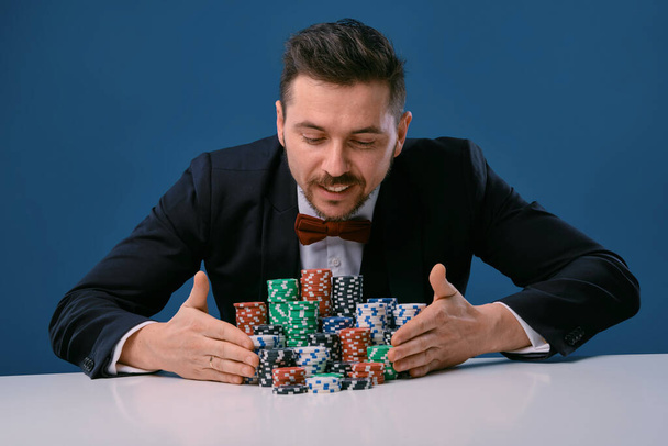 Man in black suit is sitting at white table with colored stacks of chips on it, posing on blue studio background. Gambling, poker, casino. Close-up. - Foto, afbeelding