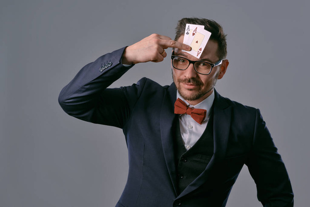 Man in black classic suit, red bow-tie, glases is showing two playing cards, posing on gray studio background. Gambling, poker, casino. Close-up. - Fotografie, Obrázek