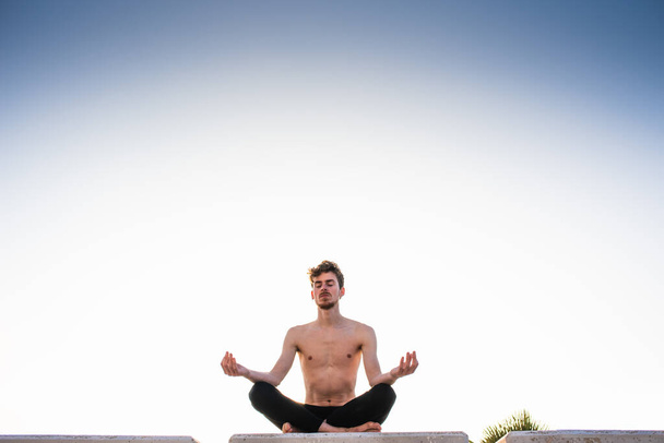 Jonge yoga beoefenaar traint haar poses, benen gekruist zittend op een houten vloer park in de stad in de schemering, lotus poseren. - Foto, afbeelding
