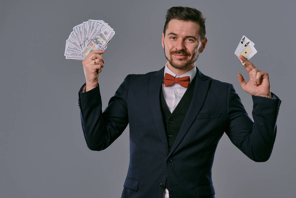 Man in black classic suit and red bow-tie showing two playing cards and cash, posing on gray studio background. Gambling, poker, casino. Close-up. - Fotó, kép