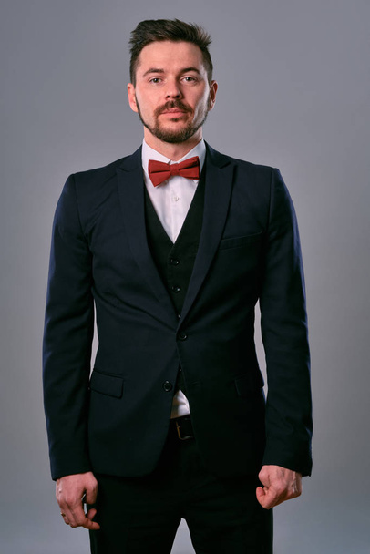 Young business man in classic black suit, white shirt and red bow-tie. Posing against a gray studio background. Mock up, copy space. Close-up. - Φωτογραφία, εικόνα