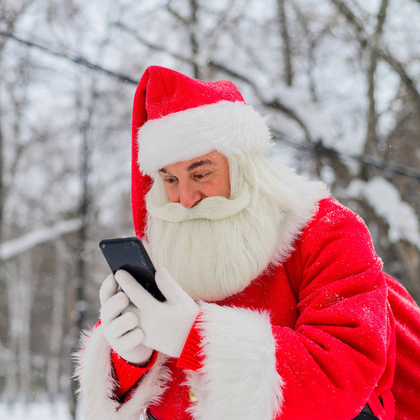 Verrast dat de kerstman buiten op de Noordpool een mobieltje gebruikt. De Kerstman staat in het park in de winter en roept de smartphone aan. Sneeuw in december. - Foto, afbeelding