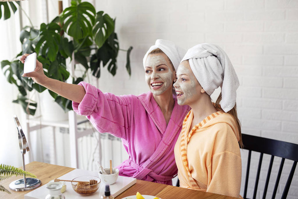 Mother and her daughter taking selfie by phone with facial mask and towel on head - 写真・画像