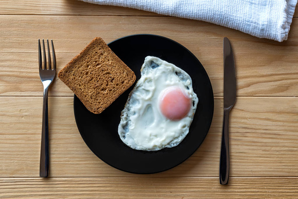 Жареное яйцо закрывается в черной тарелке на деревянном столе с хлебом и посудой. Завтрак. Солнечная сторона яичницы
 - Фото, изображение