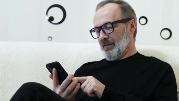 Middle-aged hipster man with a gray beard and glasses use mobile phone at home - Filmati, video