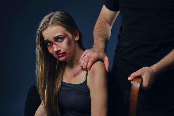 Strong man put his hand on female shoulder, showing belt. Victim with bruises on face sitting nearby. Blue background. Domestic violence. Close-up. - Φωτογραφία, εικόνα