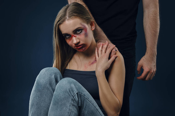 Strong man put his hand on female shoulder. Scared victim with bruises on face sitting nearby. Blue background. Domestic violence, abuse. Close-up. - Fotografie, Obrázek