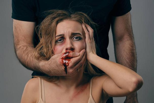 Een sterke man bedekte de mond van een bange vrouw met blauwe plekken op haar gezicht. Het bloed stroomt door zijn arm. Grijze achtergrond. Huiselijk geweld. Close-up. - Foto, afbeelding