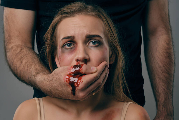 Strong man covered mouth of scared female with bruises on her face. Blood from it flows down his arm. Gray background. Domestic violence. Close-up. - Photo, Image